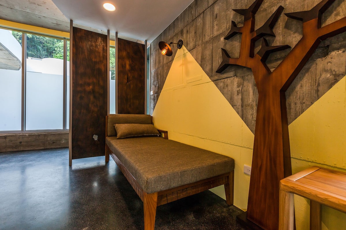 A room with a bed, a wooden bench, and a wall with a tree-like design.