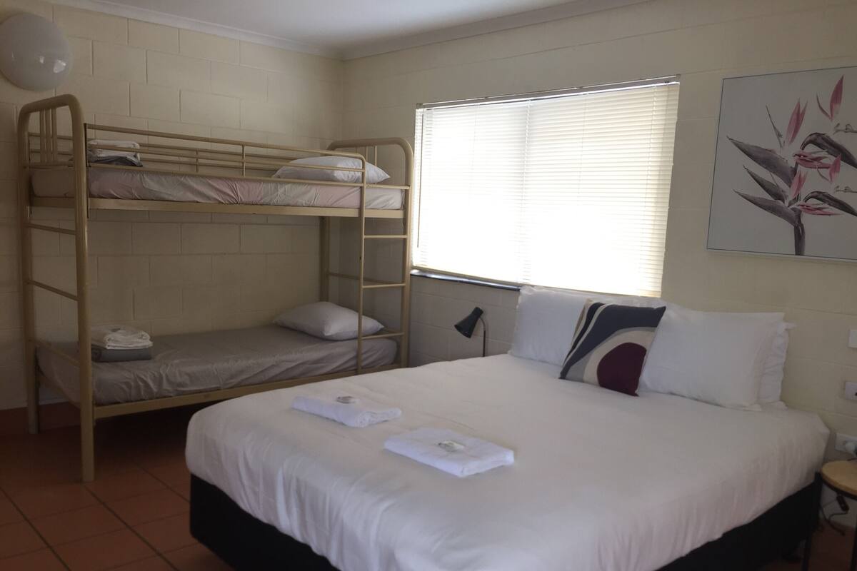 A room with a bunk bed, a single bed, a small table, and a window with blinds.