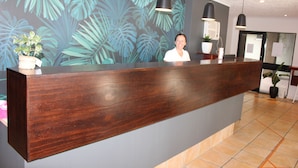 Un mostrador de recepción con acabado de madera oscura, una planta en maceta grande a la izquierda y una pared con estampado de hojas tropicales.