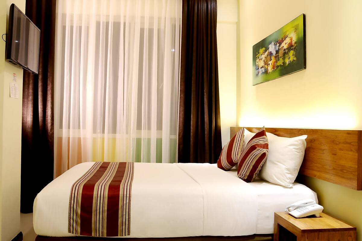 A hotel room with a bed, a bedside table with a phone, a painting on the wall, and curtains.