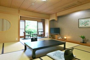 A traditional Japanese room with tatami flooring, a low table, a TV, and a view of greenery outside.