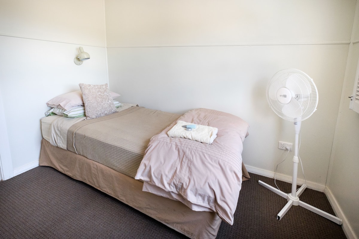 A small, neatly made bed with a beige and pink bedding set, a white standing fan, and a wall-mounted light fixture.