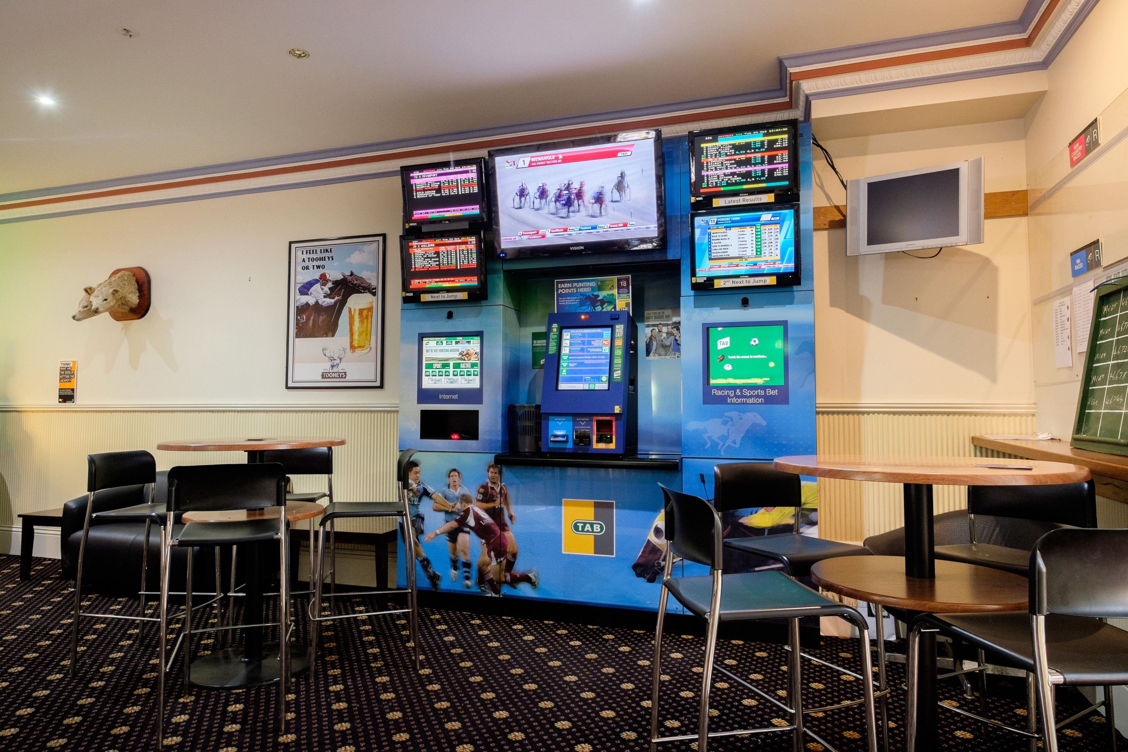 A betting counter with multiple screens displaying sports events, a mounted animal head, and several chairs and tables.