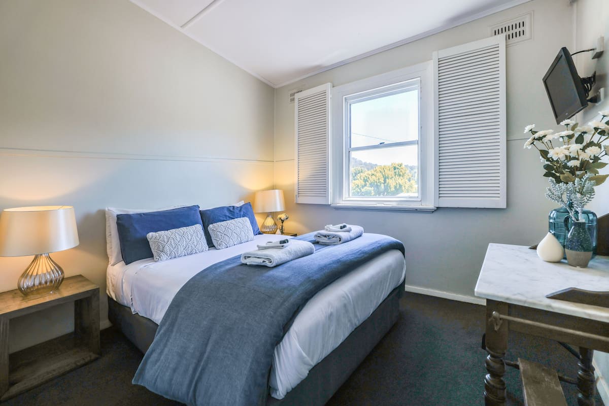 A bedroom with a bed, bedside lamps, a desk with a vase, and a window with shutters.