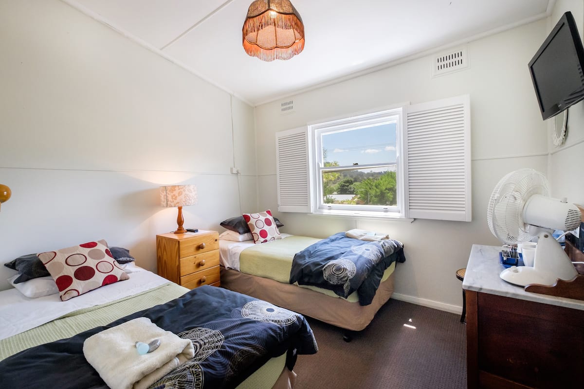 A bedroom with two beds, a desk, a chair, a lamp, a TV, and a window with shutters.