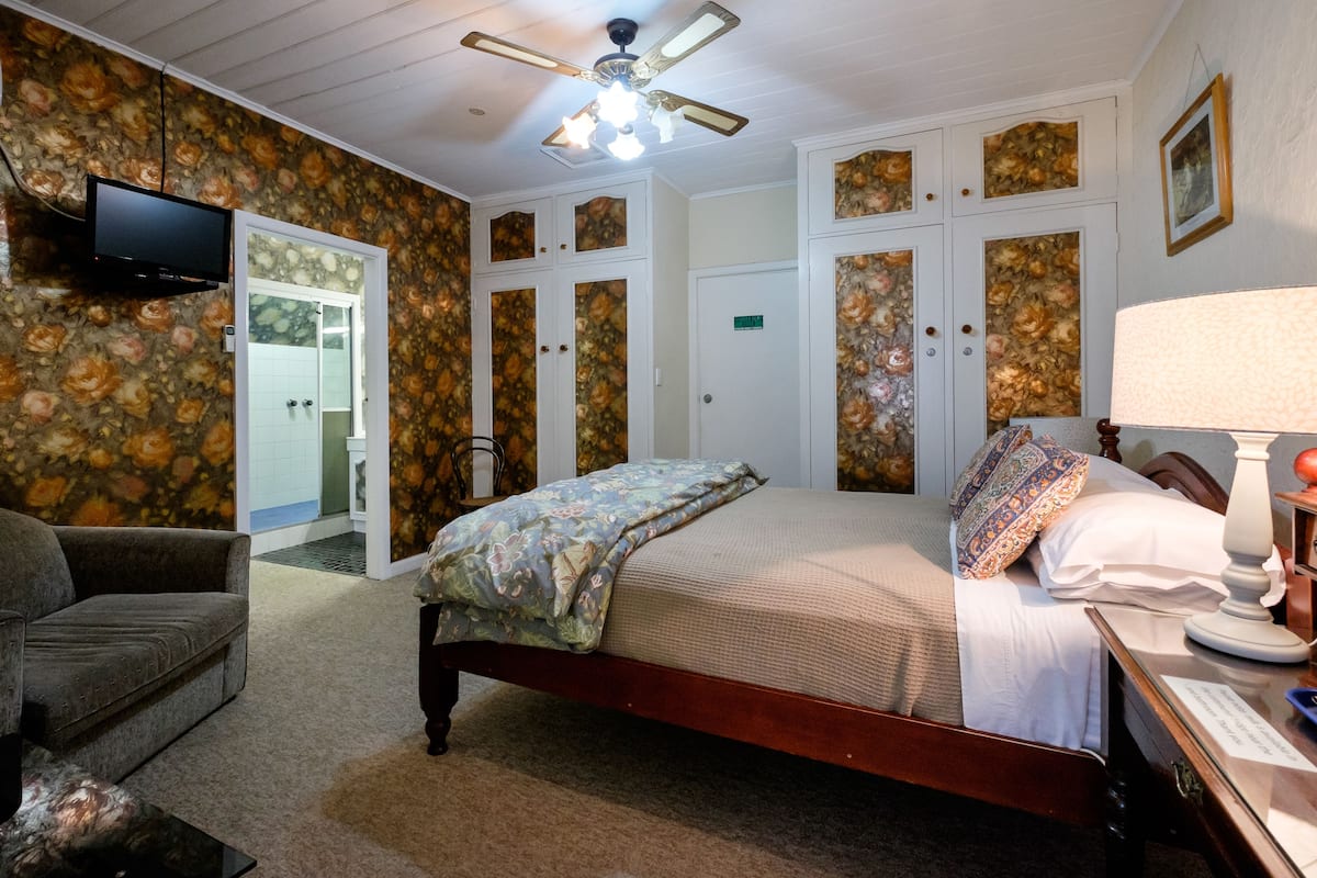 A bedroom with a bed, a chair, a desk with a lamp, a television mounted on the wall, and a ceiling fan.