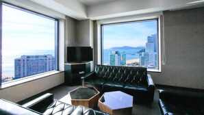 A modern living room with a leather sofa, a coffee table, and a flat-screen TV. Large windows offer a view of the city skyline and the ocean. - Benikea Hotel Haeundae (Busan)