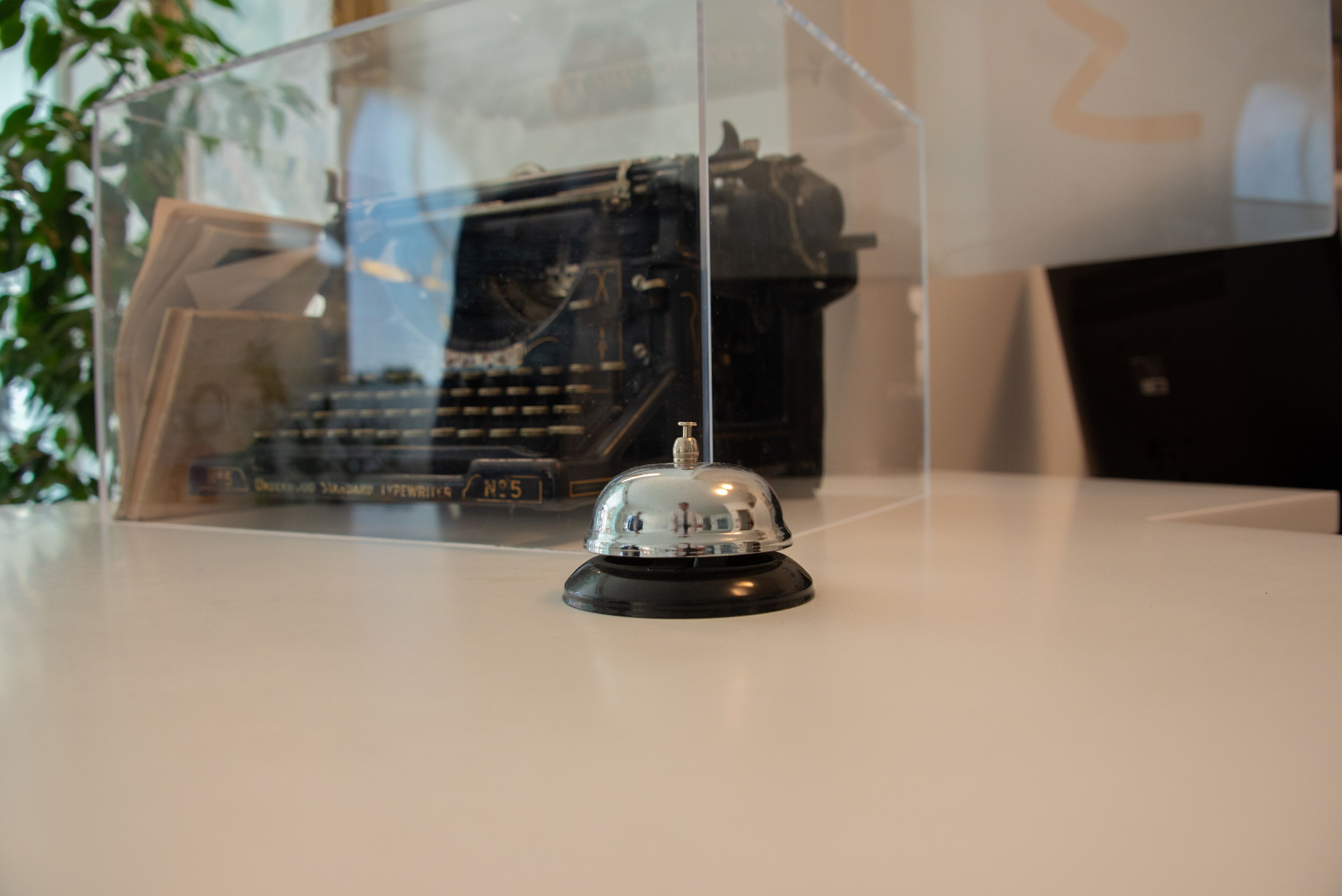 A vintage typewriter encased in a clear display, with a bell on top, and a partial view of a plant in the background.