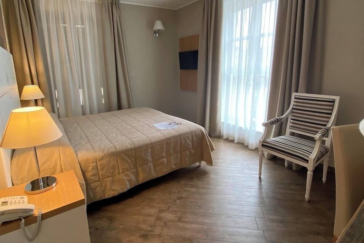 A hotel room with a bed, a desk with a lamp, a chair, and a window with curtains.