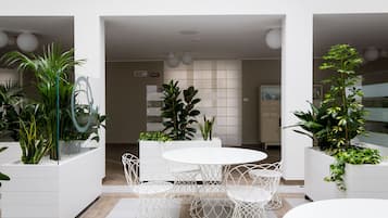 Un espacio interior moderno con una mesa redonda y sillas de estructura metálica blanca, rodeado de macetas blancas con plantas verdes.