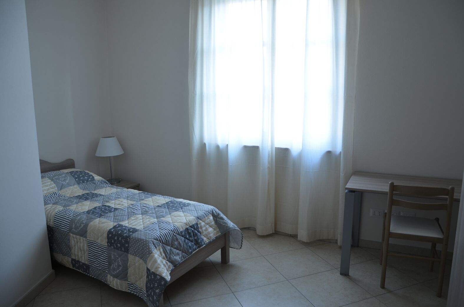 Une chambre à coucher avec un lit, un bureau, une chaise, une lampe et une fenêtre avec des rideaux.