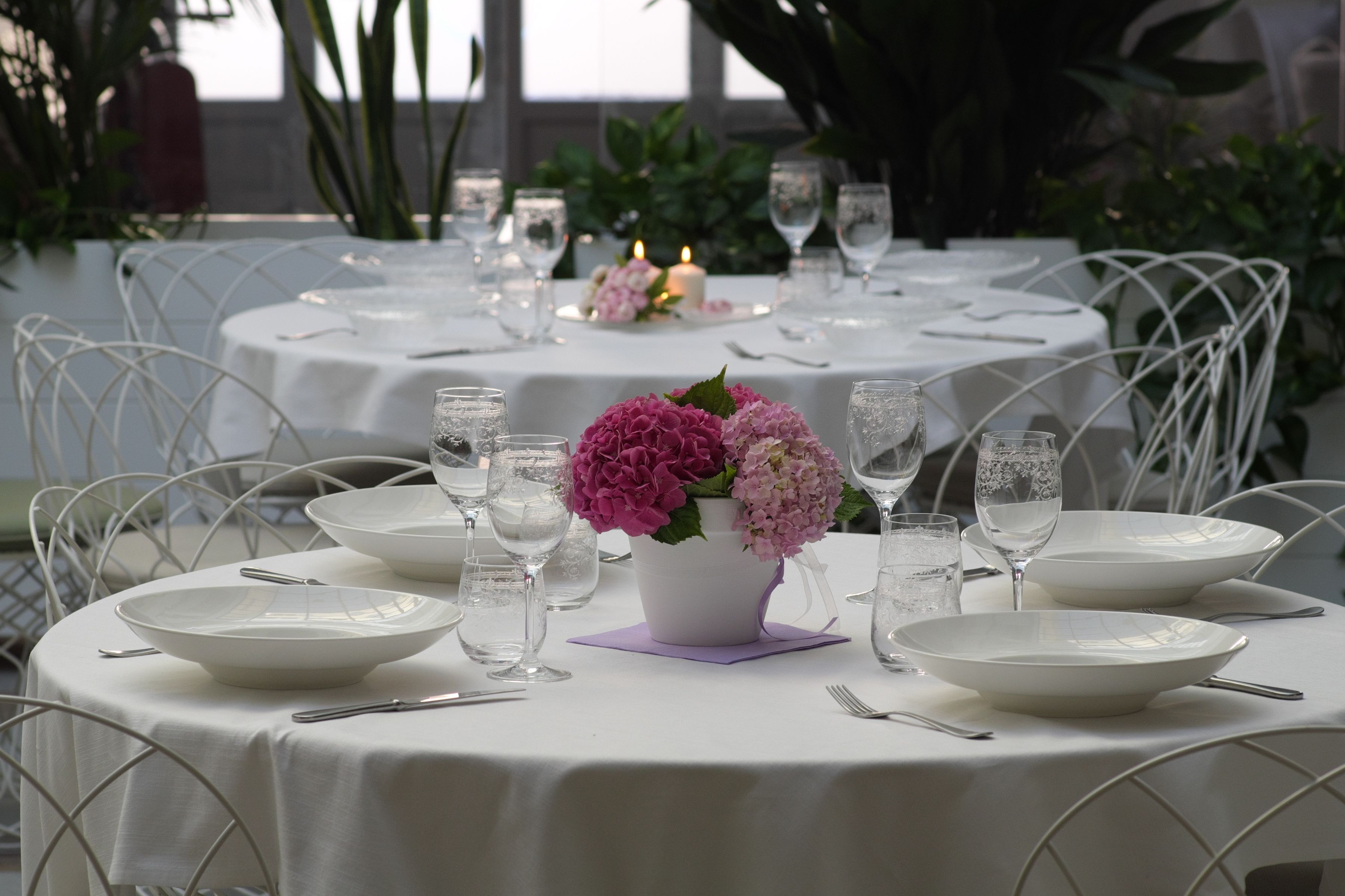 A table set for a meal with white tablecloths, clear glassware, and a vase with pink flowers.