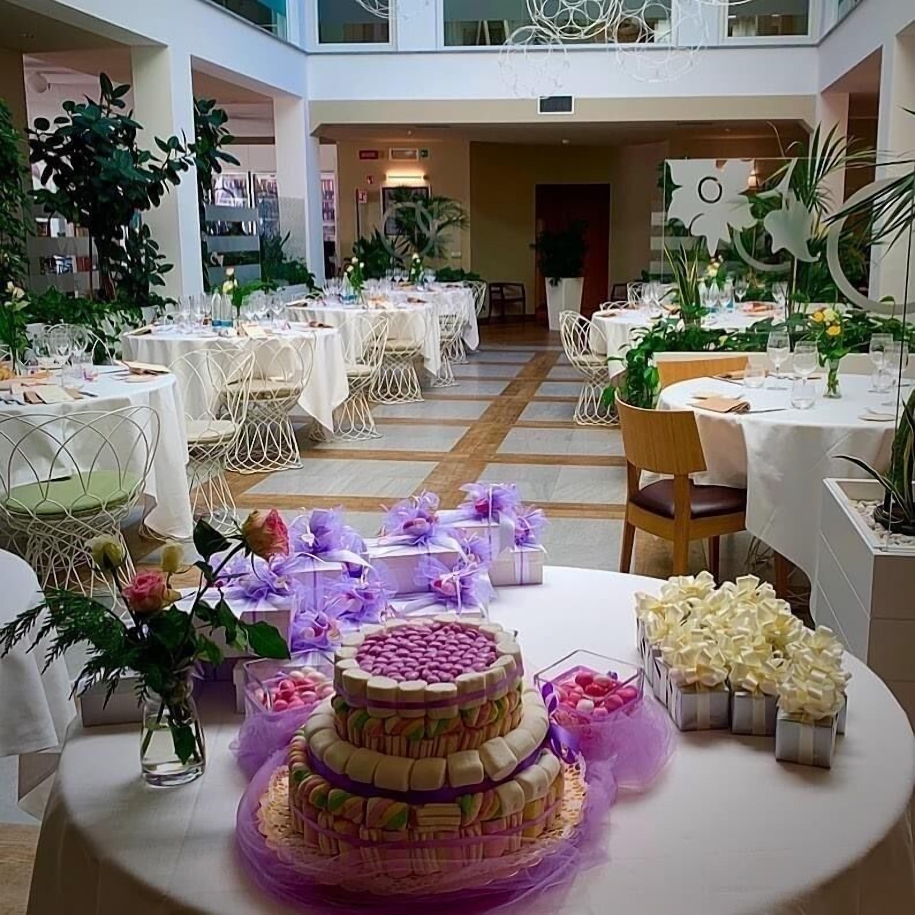A spacious indoor venue with multiple round tables set for dining, surrounded by potted plants and decorative elements.