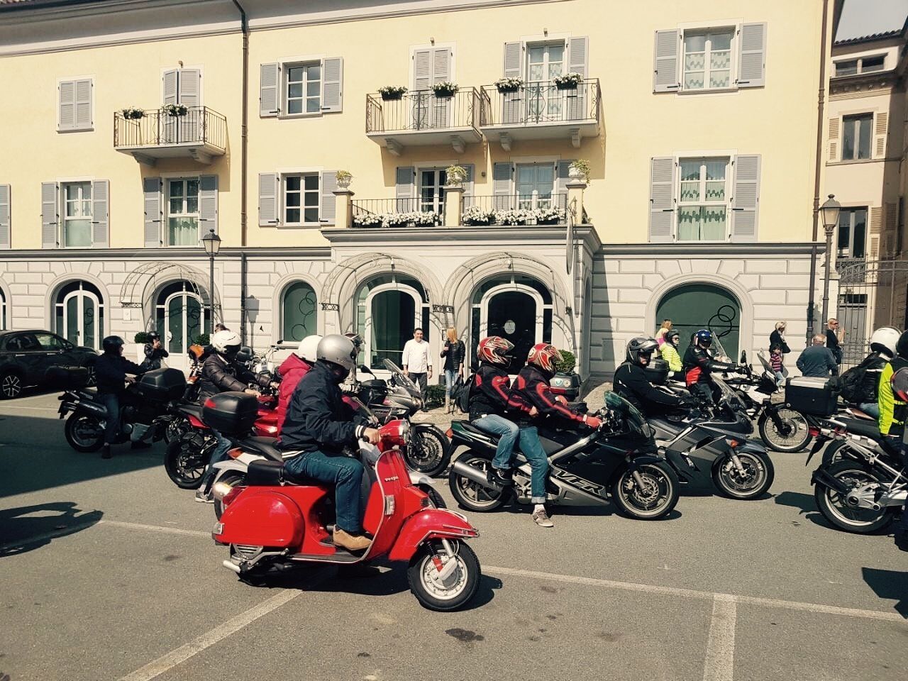 A group of motorcyclists parked in front of a building with balconies and arched windows.