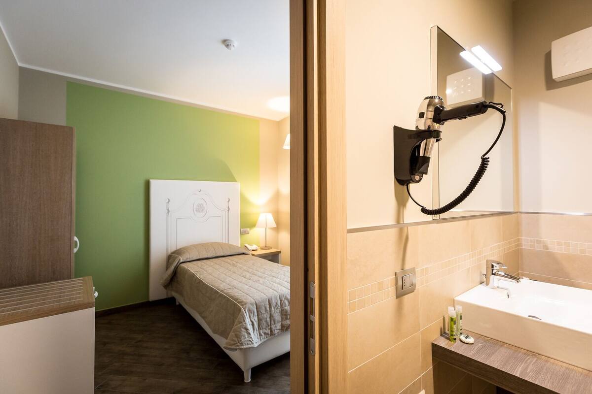 A hotel room with a bed, a bedside lamp, a hairdryer, and a bathroom with a sink and mirror.
