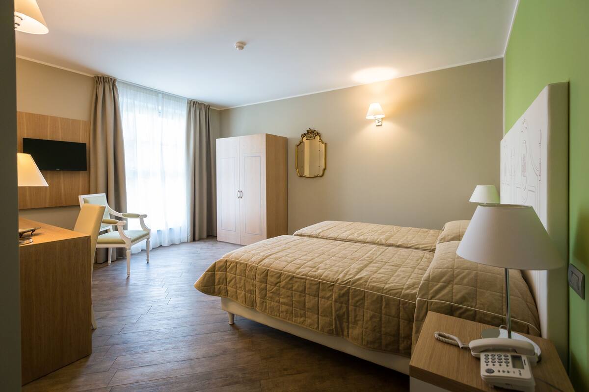 A hotel room with a bed, bedside table with a lamp, a desk with a chair, a TV mounted on the wall, and a wardrobe.