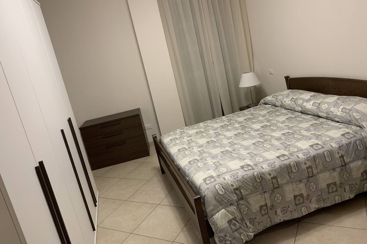 A bedroom with a bed, a nightstand, a lamp, a wardrobe, and a window with curtains.
