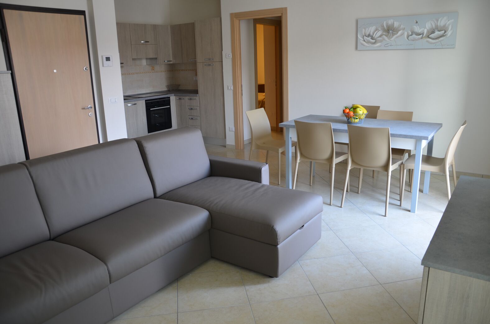 A modern living room with a grey sectional sofa, a dining area with a white table and chairs, and a kitchen in the background.