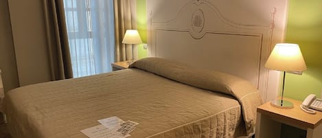 Una habitación de hotel con una cama, dos lámparas de noche, una mesita de noche con teléfono y una ventana con cortinas.