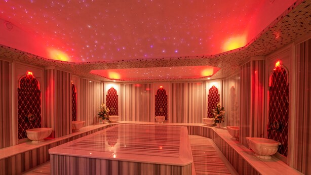 A luxurious spa room with a central marble pool, ornate red and white decor, and a starry ceiling effect.