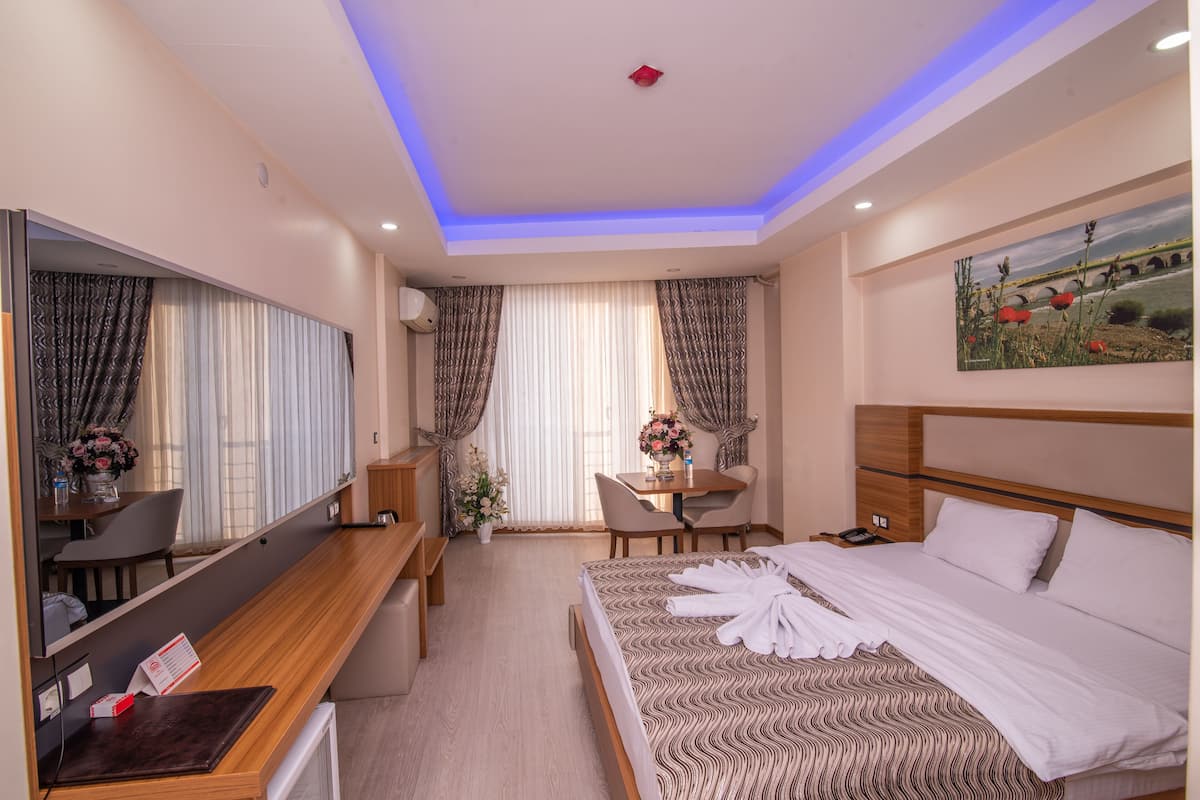 A hotel room with a bed, a desk, a chair, a table, a mirror, and a painting on the wall.