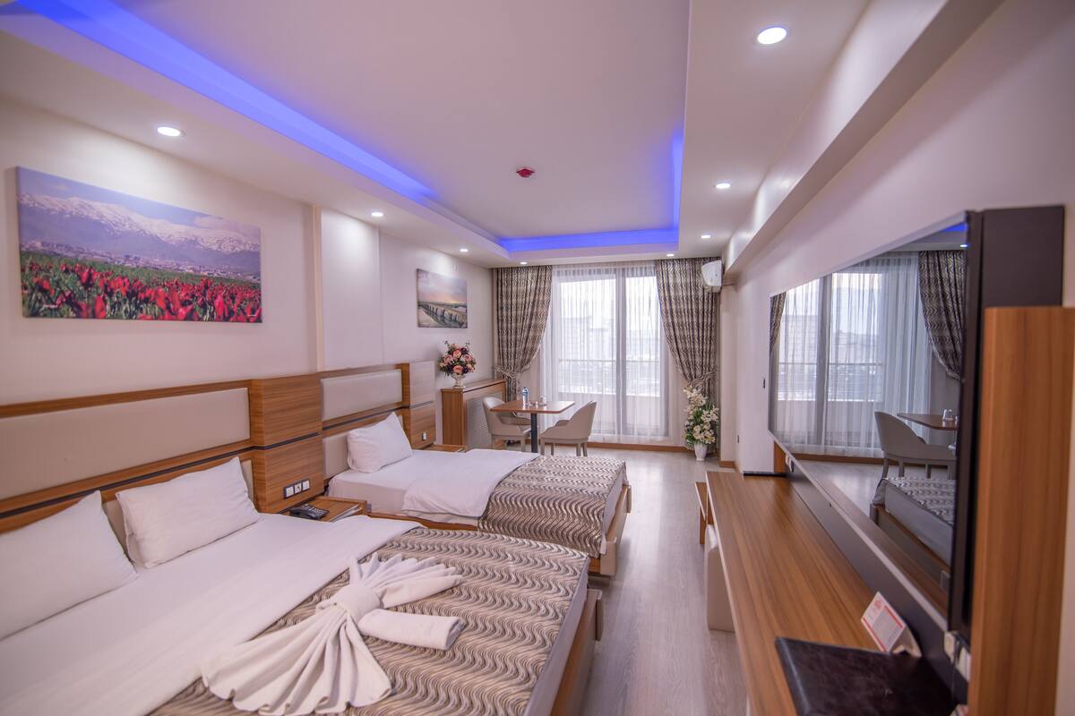 A hotel room with two beds, a desk, a TV, and a large window with curtains.