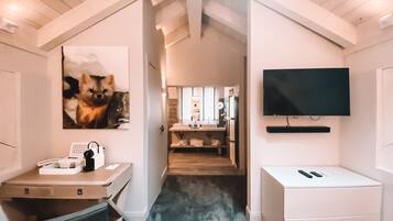 Una habitación moderna y bien iluminada, con escritorio, silla y televisor montado. Un pasillo conduce a un baño con lavabo y espejo.