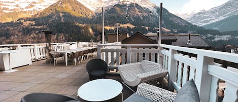 A terrace with outdoor seating, a view of snow-capped mountains, and a clear sky.