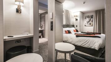 Una habitación de hotel moderna con una cama grande, una mesita redonda pequeña, un espejo y una lámpara de pared.