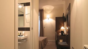 A hotel hallway with a bathroom on the left, a bed in the center, and a desk with a lamp on the right. - Andrea Doria Resort (Rome)
