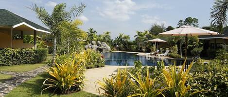 A pool area with lounge chairs and umbrellas, surrounded by tropical plants and a stone pathway.