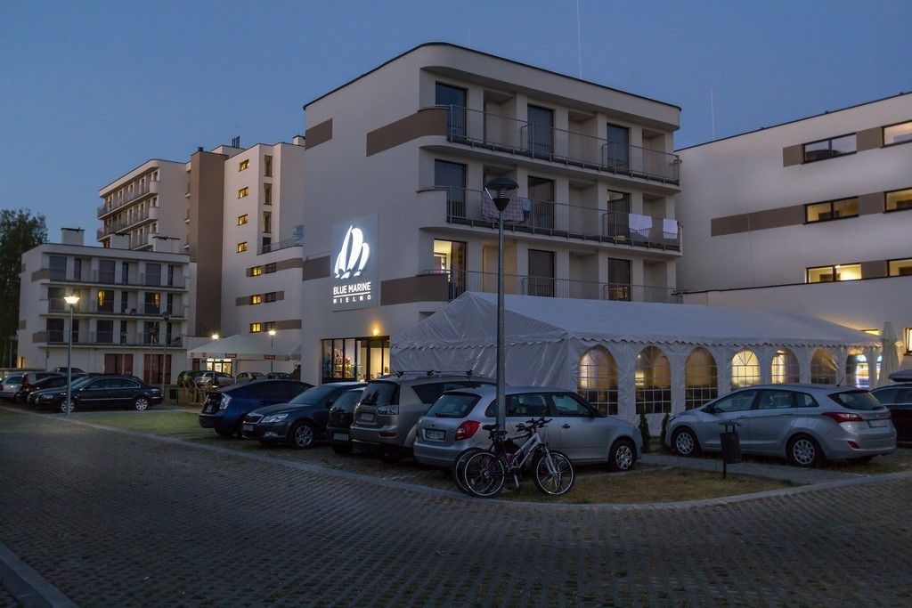 Un hotel con fachada moderna, un letrero visible que dice 'Blue Marine', y un área exterior cubierta con coches y bicicletas estacionados.