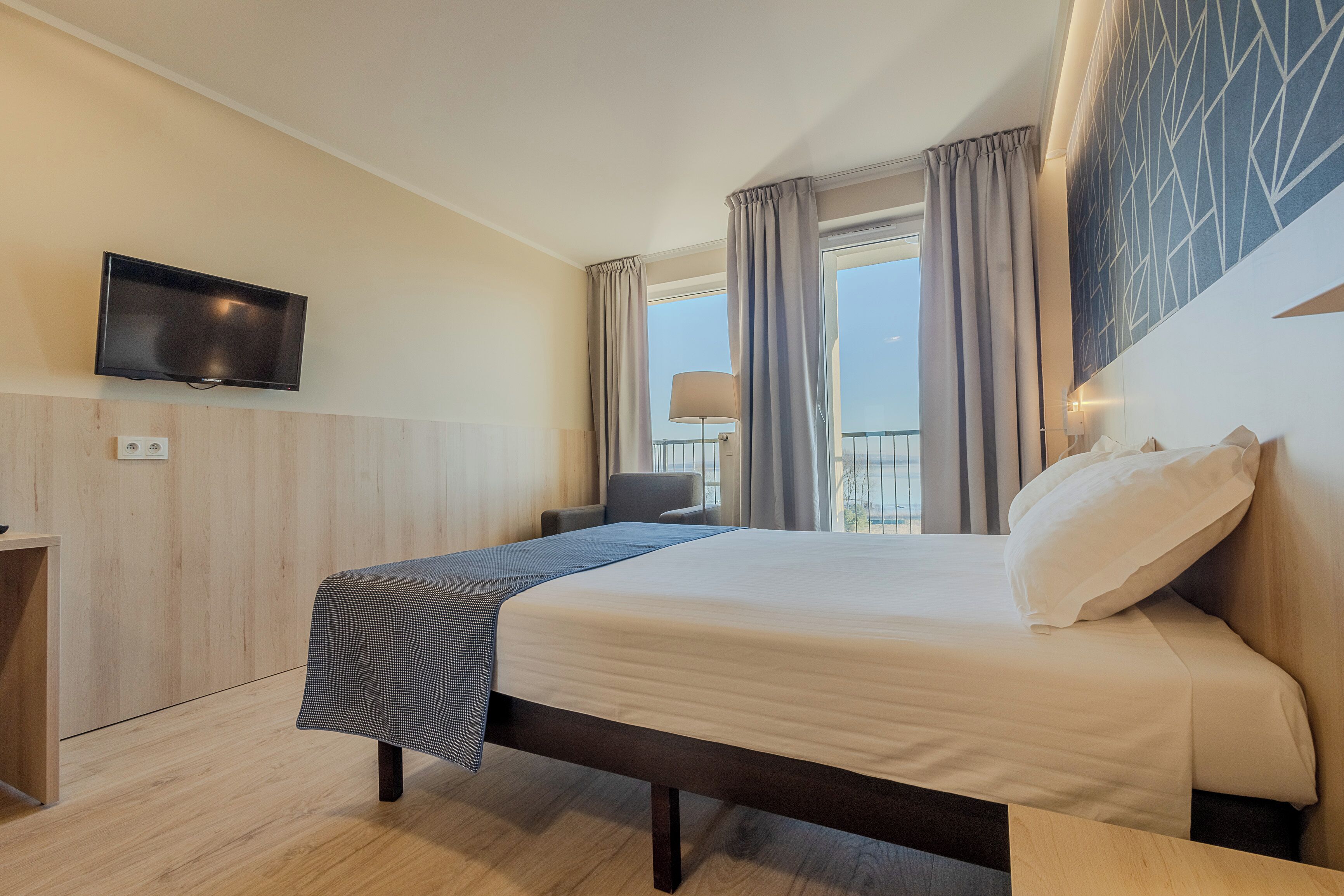 Habitación de hotel con cama, televisor montado en la pared, ventana con cortinas y vistas al exterior.