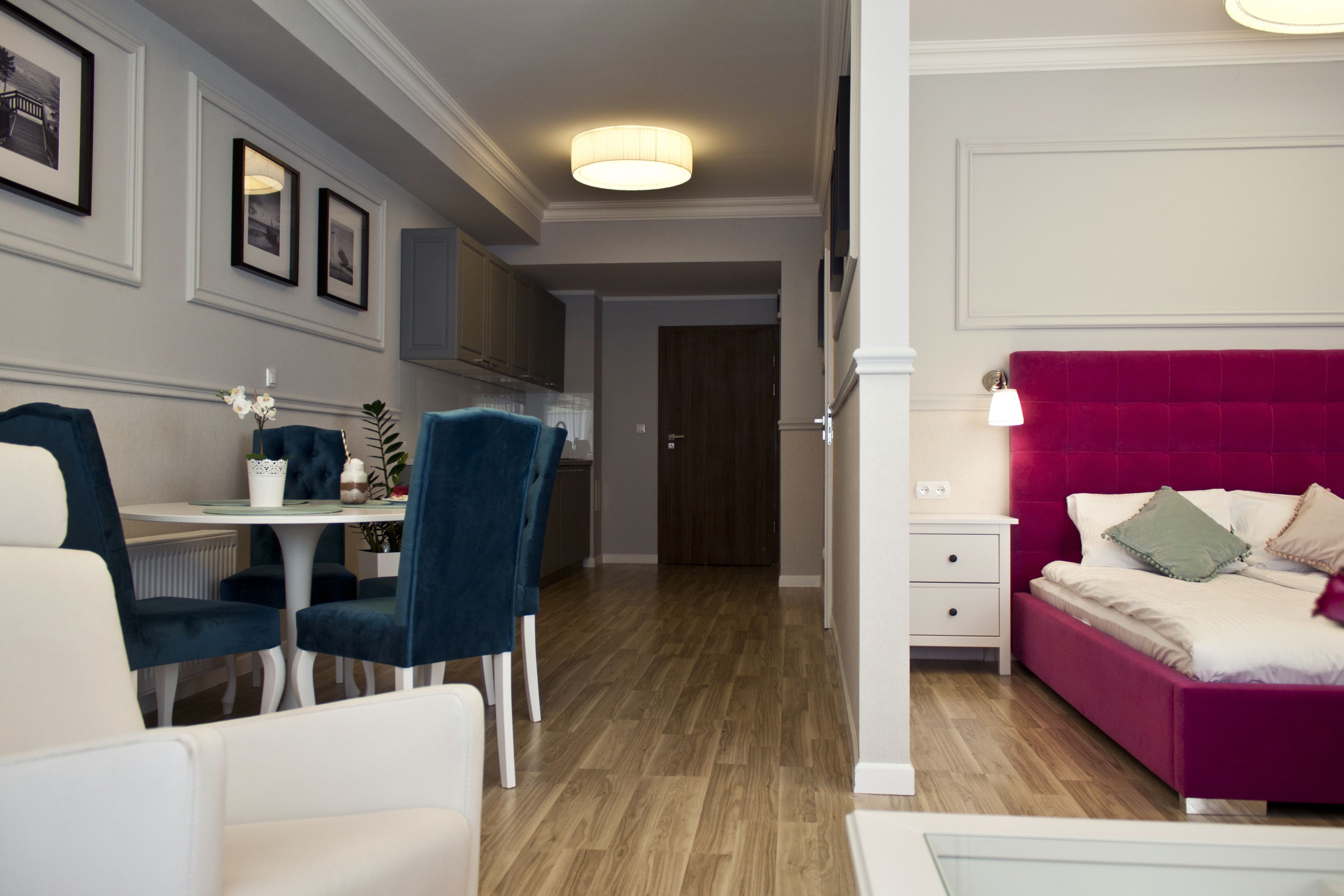 A modern hotel room with a dining area, a bed with a red headboard, and a hallway leading to a door.