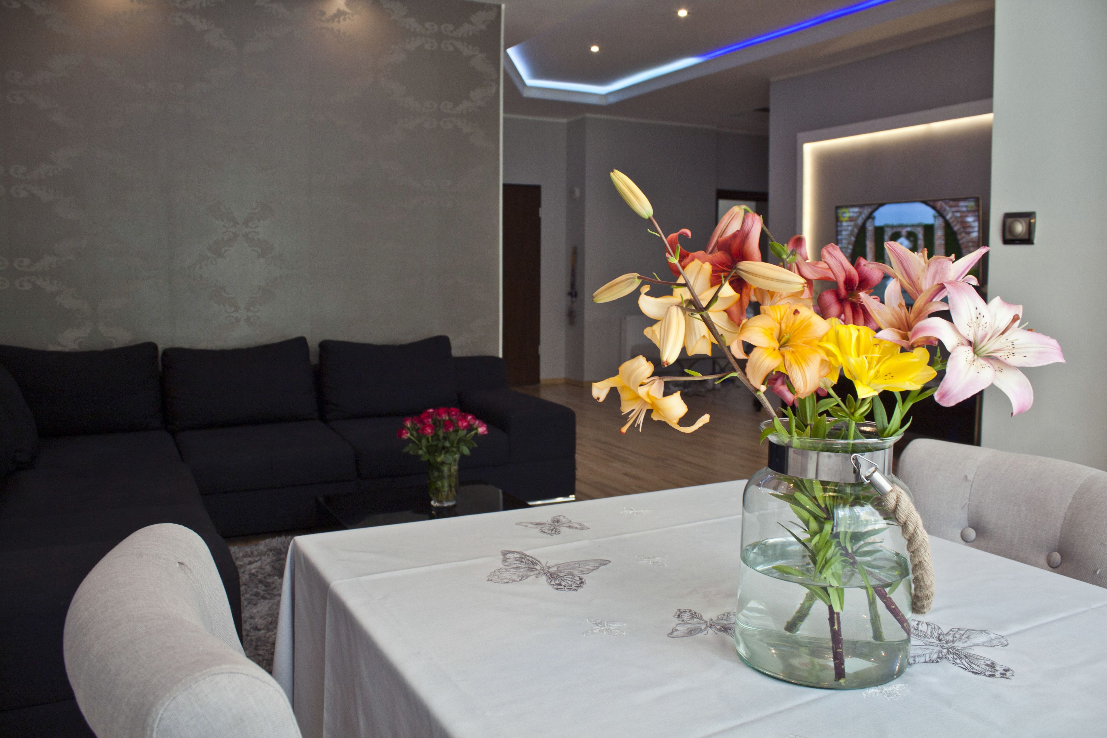 A modern living room with a black sofa, a glass vase with flowers on a table, and a decorative butterfly tablecloth.