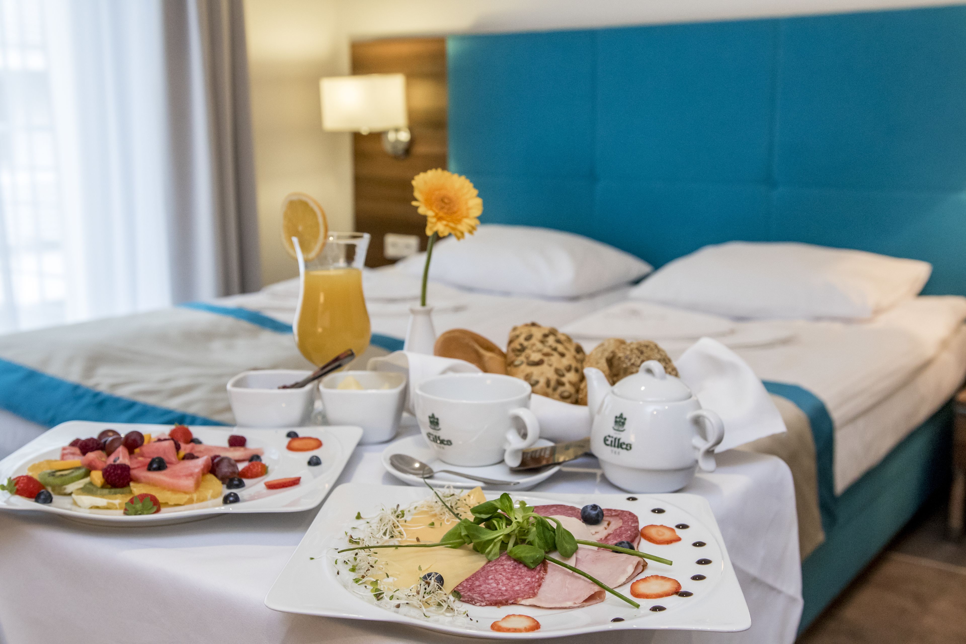 A neatly arranged breakfast table with plates of fruit, cheese, and meats, accompanied by coffee cups and a teapot, set against a bed with a blue headboard.