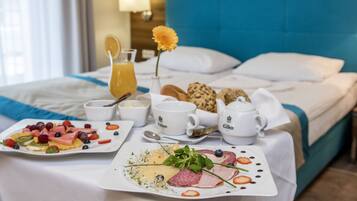 A neatly arranged breakfast table with plates of fruit, cheese, and meats, accompanied by coffee cups and a teapot, set against a bed with a blue headboard.