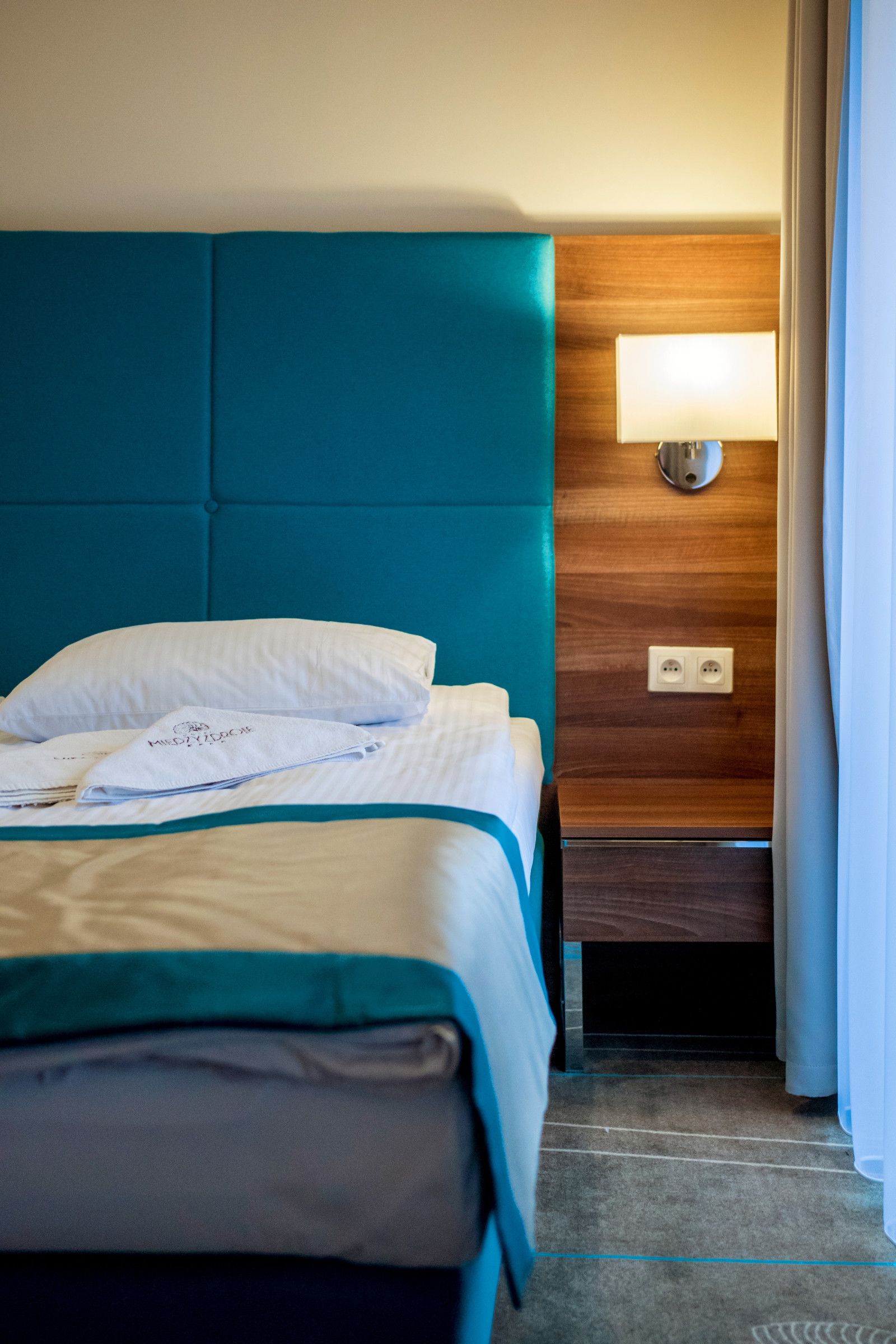 Een hotelkamer met een blauwgroen bekleden hoofdeinde, een houten nachtkastje met een lamp en een bed met witte lakens.