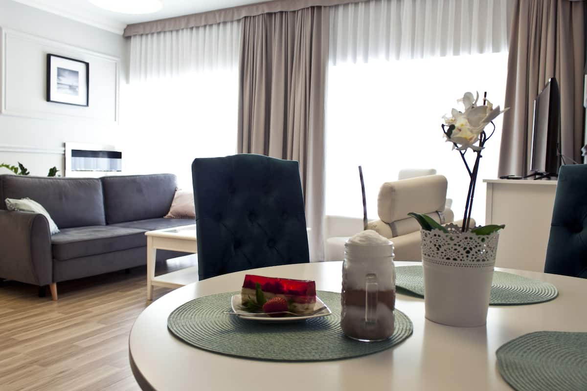 A modern living room with a grey sofa, a dark blue armchair, and a white coffee table. There is a small white table with a plate of food and a jar of coffee. A potted plant with white flowers is on the coffee table.