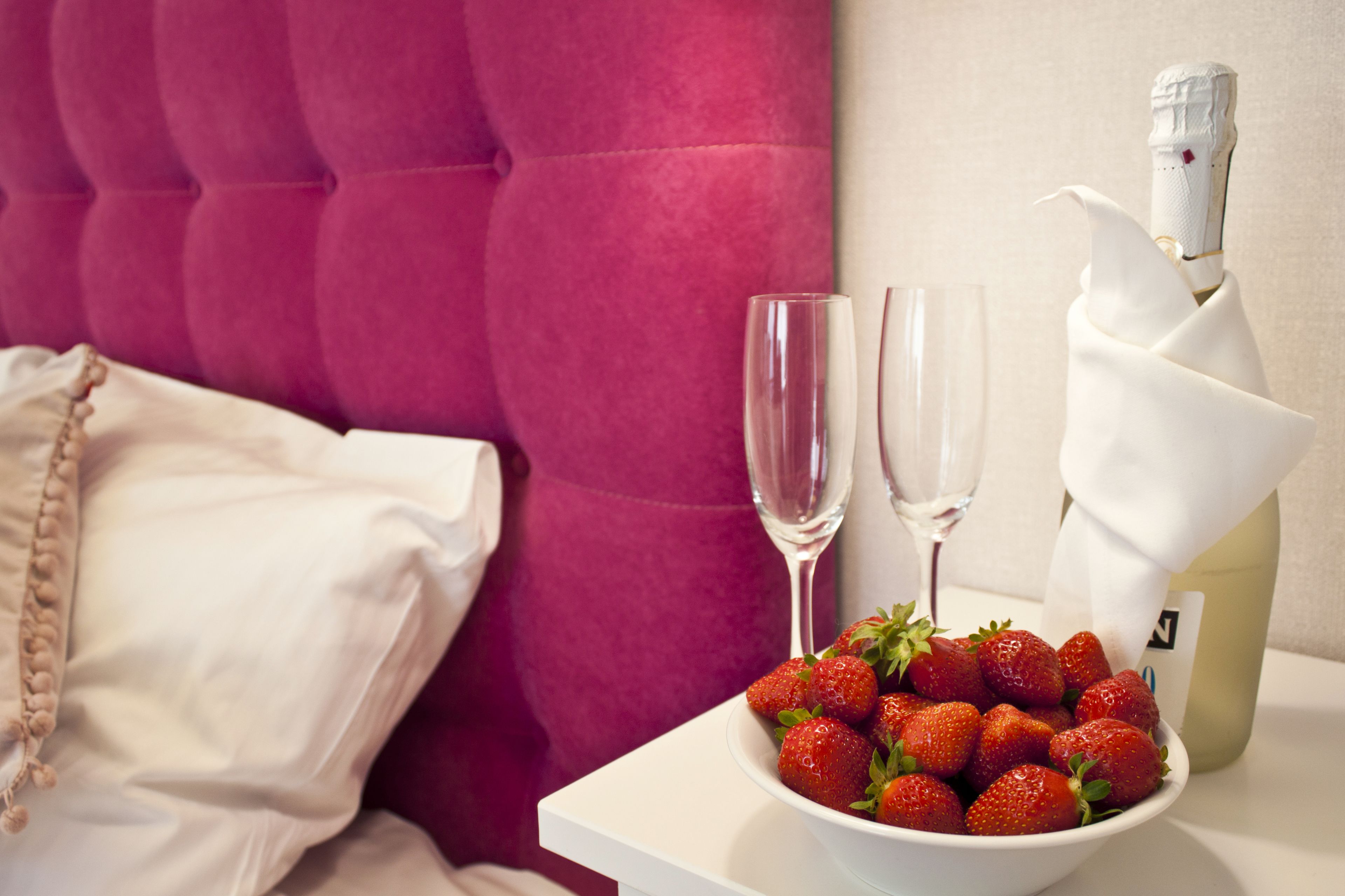 Un lit avec une tête de lit rose, un oreiller blanc, un bol de fraises, deux coupes à champagne et une bouteille de champagne.