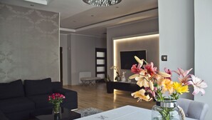 A modern living room with a black sofa, a coffee table, and a vase of flowers. There is a flat-screen TV mounted on the wall and a wooden floor. - Aparthotel Miedzyzdroje (Miedzyzdroje)