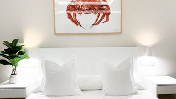 A neatly made bed with white bedding and pillows, a framed artwork of a crab on the wall, and a potted plant on the nightstand.