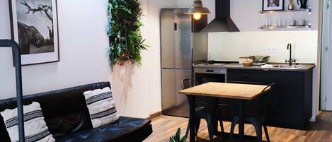 A modern living space with a black leather sofa, a wooden dining table, and a kitchen area with a stainless steel refrigerator and black cabinets.