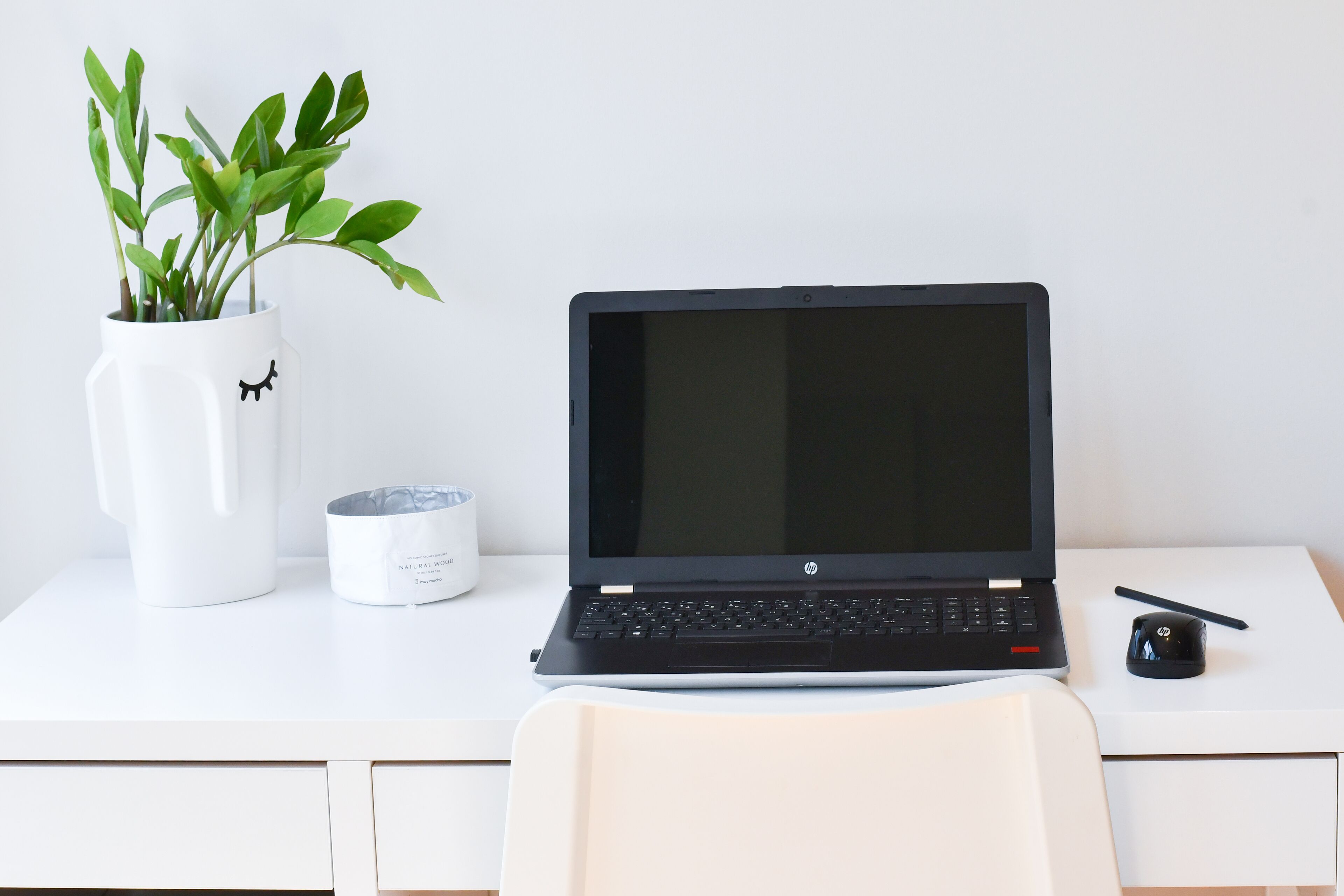 Una computadora portátil sobre un escritorio blanco, con una planta en maceta, una taza blanca y un ratón negro.