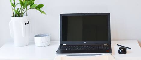 Una computadora portátil sobre un escritorio blanco, con una planta en maceta, una taza blanca y un ratón negro.