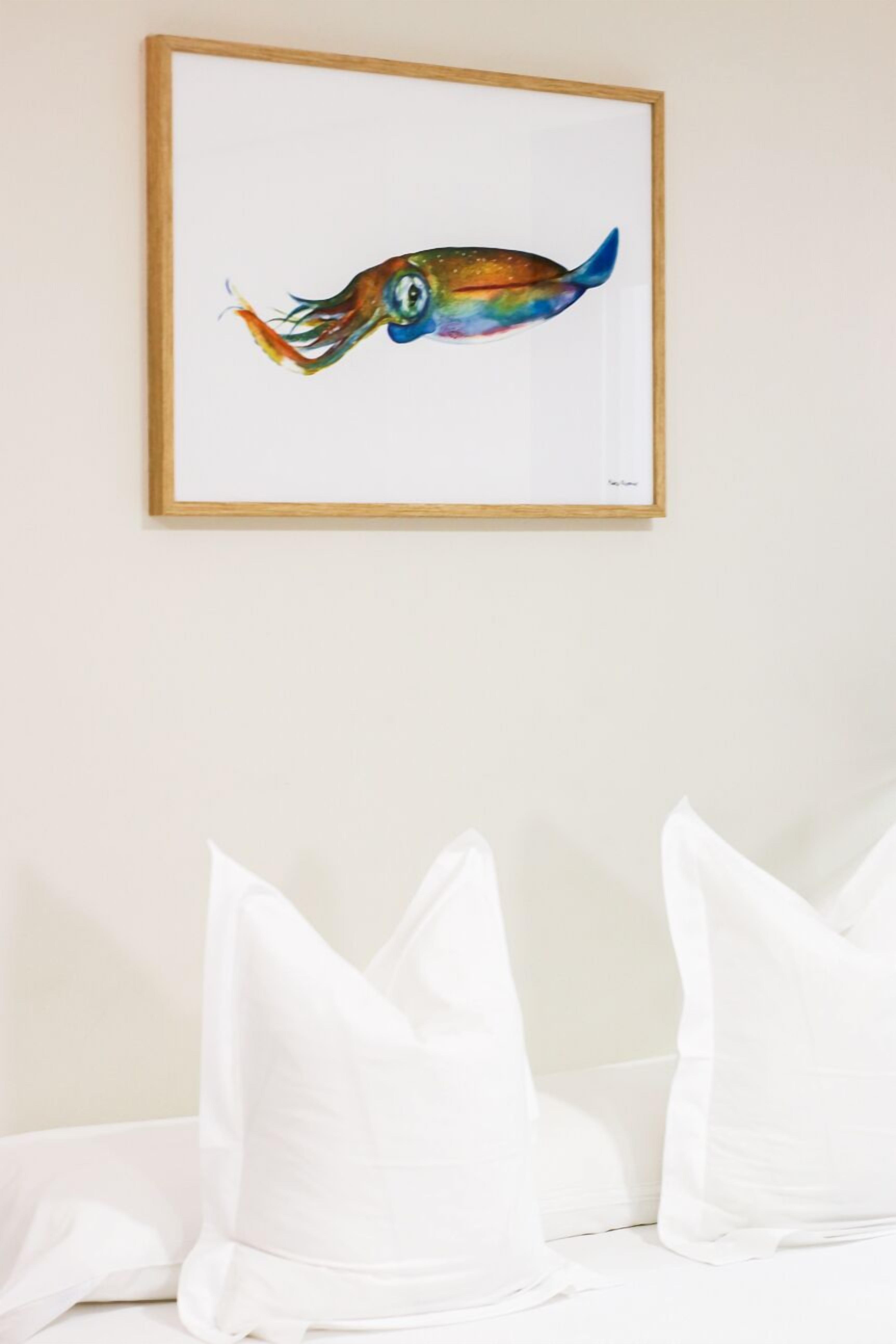 Ein gerahmtes Bild von einem bunten Fisch hängt über einem Bett mit weißen Kissen an der Wand.