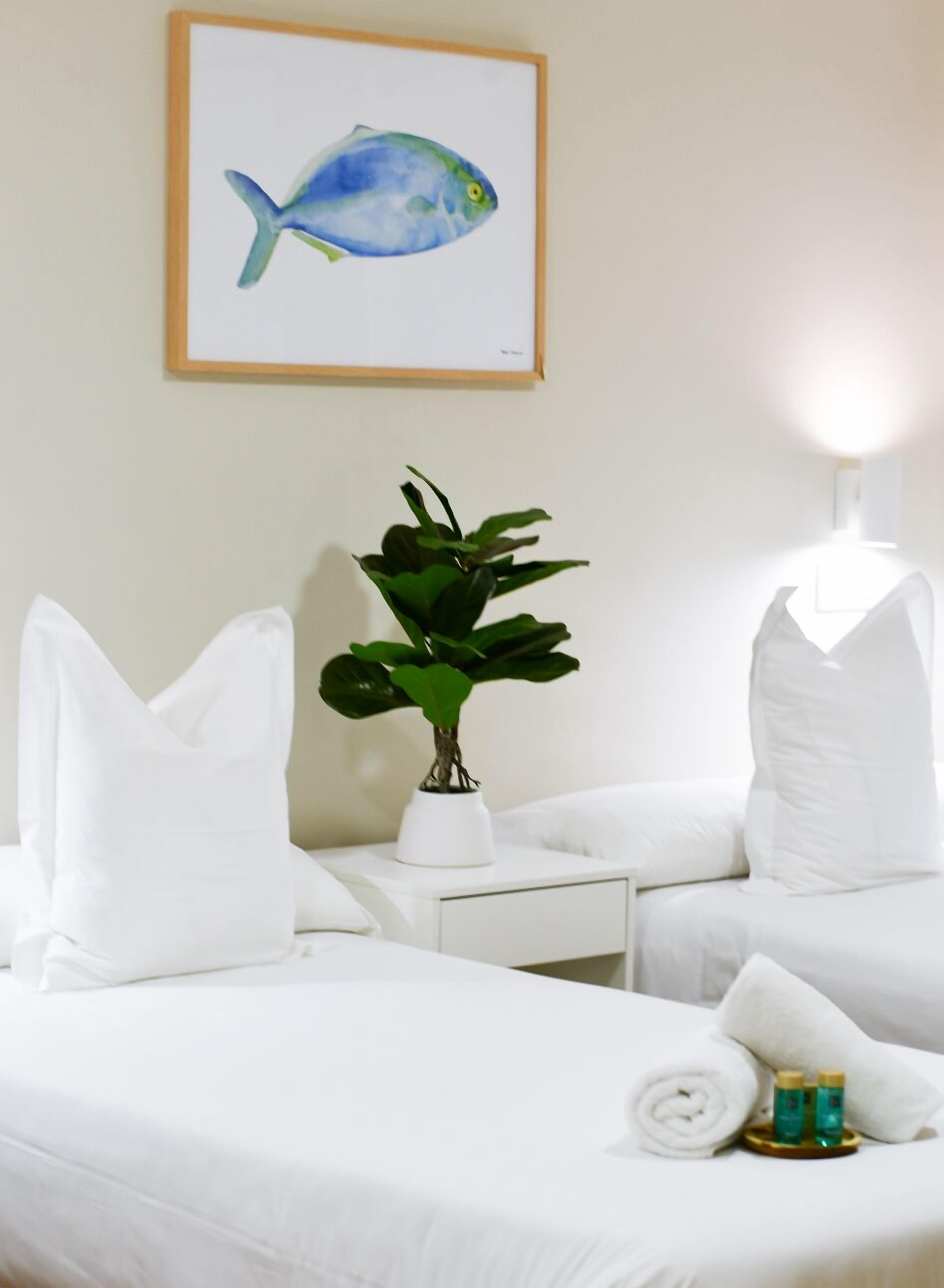 A hotel room with two beds, white bedding, a potted plant, and a framed fish painting on the wall.