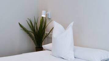 Un lit bien fait, avec du linge de lit blanc, une fougère en pot et un luminaire fixé au mur.