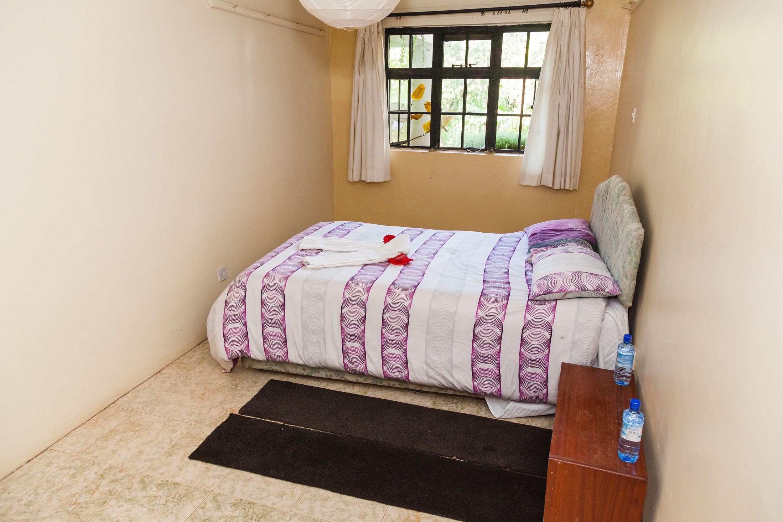 A small bedroom with a bed, a wooden nightstand, a window with curtains, and a black mat on the floor.