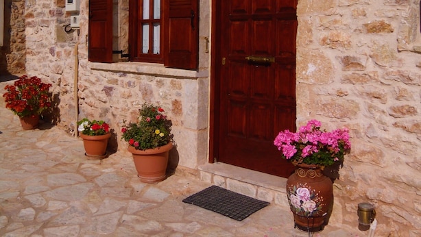 A stone building with a red door and window shutters, potted plants, and a metal grate on the ground. - Arhontiko Kordopati Traditional Guesthouse (Tripoli)